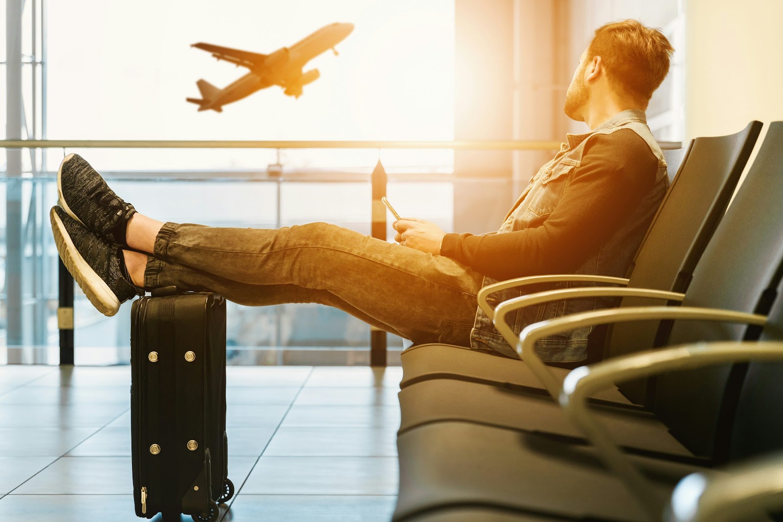 Man sat in an airport, resting his legs on a suitcase, while watching a plane take off during sunset.